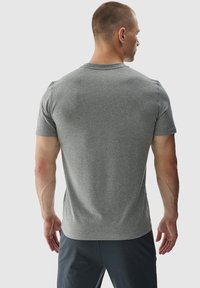 Hombre con el cabello cortado que lleva una camiseta gris de manga corta y pantalones oscuros, de pie de espaldas contra un fondo sencillo.