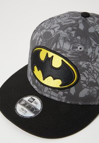 Grau Baseballcap mit einem schwarzen Schirm, ausgestattet mit einem gelben und schwarzen Batman-Logo-Patch. Strukturiertes Material mit einem Allover-Druck-Hintergrund.