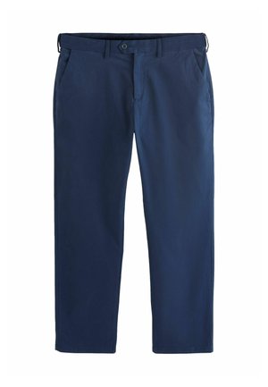 Man met marineblauwe broek, bruine leren bootschoenen en een wit overhemd met lange mouwen met dunne blauwe strepen, binnen staand.