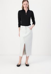Haut noir à manches longues zippé associé à une jupe en denim bleu clair présentant une fente à l'avant. La modèle porte des ballerines noires. Fond minimaliste.
