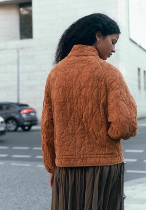 Quilted, rust-colored jacket featuring intricate embroidery, high collar, and relaxed fit. Worn over a long, pleated brown skirt.