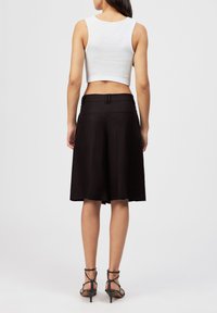 Kvinde iført sorte knælange, vidde ben-shorts, en hvid cropped tanktop og sorte hælede sandaler med stropper, stående med ryggen til.