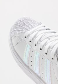 Vit lädersneaker med meshdetaljer, reflekterande ränder och texturerad tåhätta. Har vita skosnören och perforerad sida för andningsförmåga.