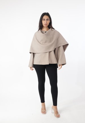 Vrouw die een beige oversized jas in cape-stijl draagt, zwarte legging en transparante sandalen met hoge hak, staand tegen een witte achtergrond.