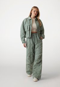Jachetă scurtă din in verde, cu două buzunare frontale, asortată cu pantaloni largi și albi și adidași albi, pe un fundal deschis.