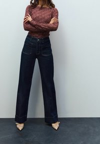 Femme debout, les bras croisés, portant un haut en dentelle bordeaux à manches longues, un jean taille haute foncé et des talons pointus beiges avec des brides.