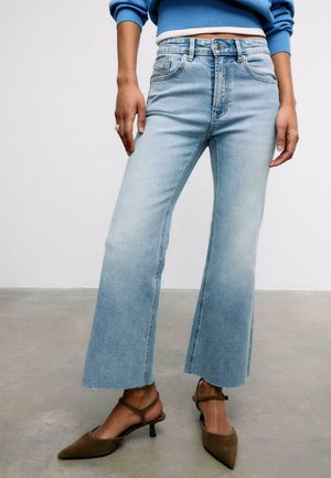 Osoba oblečená v svetlomodrých džínsach s širokými nohavicami, modrom svetri a hnedých špicatých podpätkoch stojaca na sivej podlahe pred bielou stenou.