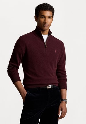 Burgundy zip-neck sweater made of soft knit fabric, with ribbed cuffs and hem. Features a small logo on the chest and paired with dark pants.