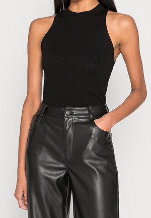 Kobieta w czarnym, prążkowanym topie bez rękawów i wysokich czarnych skórzanych spodniach z prawą ręką w kieszeni na jednolitym tle.