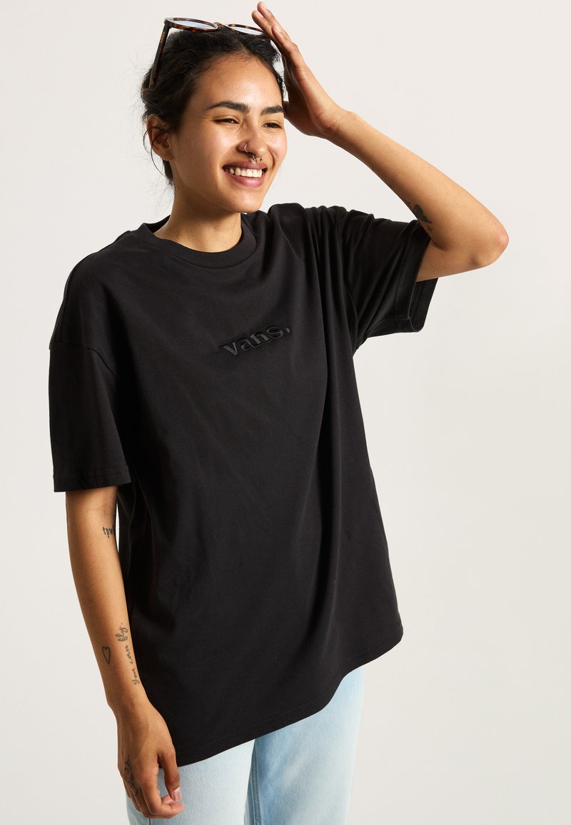 Camiseta de algodón negra con un corte relajado, que presenta un logotipo bordado de "vans." en el pecho, combinada con jeans de color azul claro.