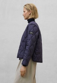 Marineblå quiltet jakke med rund hals, prydet med diagonale syninger og et mærke på venstre ærme. Båret over en sort turtleneck.