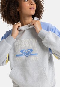 Gray sweatshirt with light blue and yellow sleeves, featuring a Roxy logo in blue and cream. Cozy, textured fabric with a kangaroo pocket.