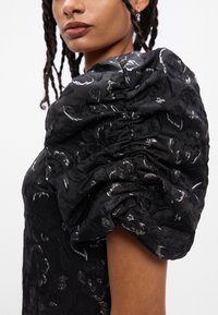 Crna haljina s napuhanim rukavima sa teksturiranim cvjetnim uzorcima i suptilnim sjajem. Krupni plan detalja rukava prikazuje skupljeno tkanje.