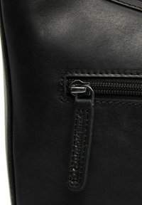 Dettaglio di una borsa in pelle nera che mostra una tasca con zip, con hardware argentato e etichetta del marchio embossata, caratterizzata da una texture liscia e cuciture.
