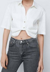 Mujer vestida con una camisa blanca de botones de manga corta atada a la cintura y vaqueros de mezclilla negros de cintura alta.