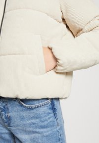 Krémová žebrovaná kordurová pufi bunda se zipem, která má boční kapsu a elastické manžety, nošená přes modré džíny.