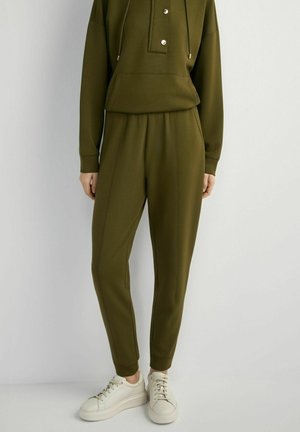Osoba ubrana w oliwkowozielone spodnie dresowe i pasującą bluzę z kapturem oraz białe trampki, stojąca na tle jasnoszarego tła.