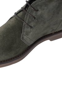 Stivale alla caviglia in suede verde con lacci marroni, superficie texture, cuciture rinforzate e suola piatta in gomma. Design semplice ed elegante.