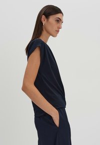 Top corto a maniche corte di colore blu navy in tessuto morbido con un davanti drappeggiato e vestibilità rilassata. Il modello è in posa di lato, mostrando le tasche e la texture liscia.