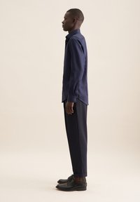Camicia a maniche lunghe blu navy in tessuto morbido, con colletto classico e polsini con bottone, abbinata a pantaloni scuri a gamba stretta e scarpe nere.