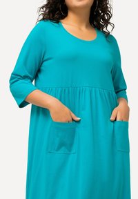 Turkusowa sukienka z okrągłym dekoltem, rękawami o długości trzech czwartek i zebraną talią. Posiada dwie kieszenie z przodu w kształcie kwadratu. Wykonana z miękkiego materiału.