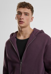 Lilla zip-up hettegenser i bomullsblanding, med frontglidelås, stor hette og kengurulomme. Laget over en svart ribbestrikket skjorte.