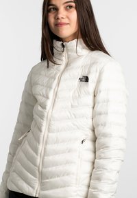 Weißer Daunenjacke mit hohem Kragen, Reißverschluss vorne und leichtem Glanz. Beinhaltet zwei zipbare Taschen und ein dezentes Logo auf der Brust.