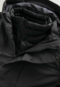 Sort jakke med lynlås, quiltet indvendig foring og tekstureret stof. Inkluderer justerbare elementer og metalliske hardware-detaljer.