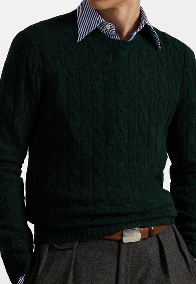 Pull en maille torsadée vert foncé superposé à une chemise à col rayée bleue et blanche. Ceinture marron avec une boucle rectangulaire en argent.