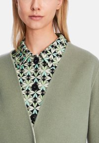 Grüne, strukturierte Strickjacke über einem gemusterten schwarzen Hemd mit mintgrünen und weißen Blumenmotiven sowie schwarzen Knöpfen.