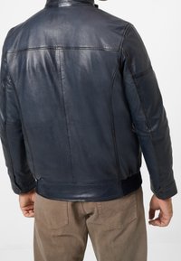 Man i mörkblå läderjacka och bruna byxor, sedd bakifrån mot en enfärgad vit bakgrund.