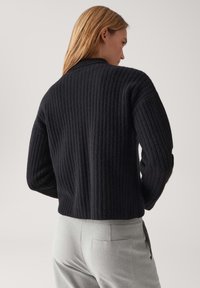 Donna con capelli castano chiaro indossa un cardigan a coste nero e pantaloni grigi chiari, mostrata di spalle contro uno sfondo semplice.