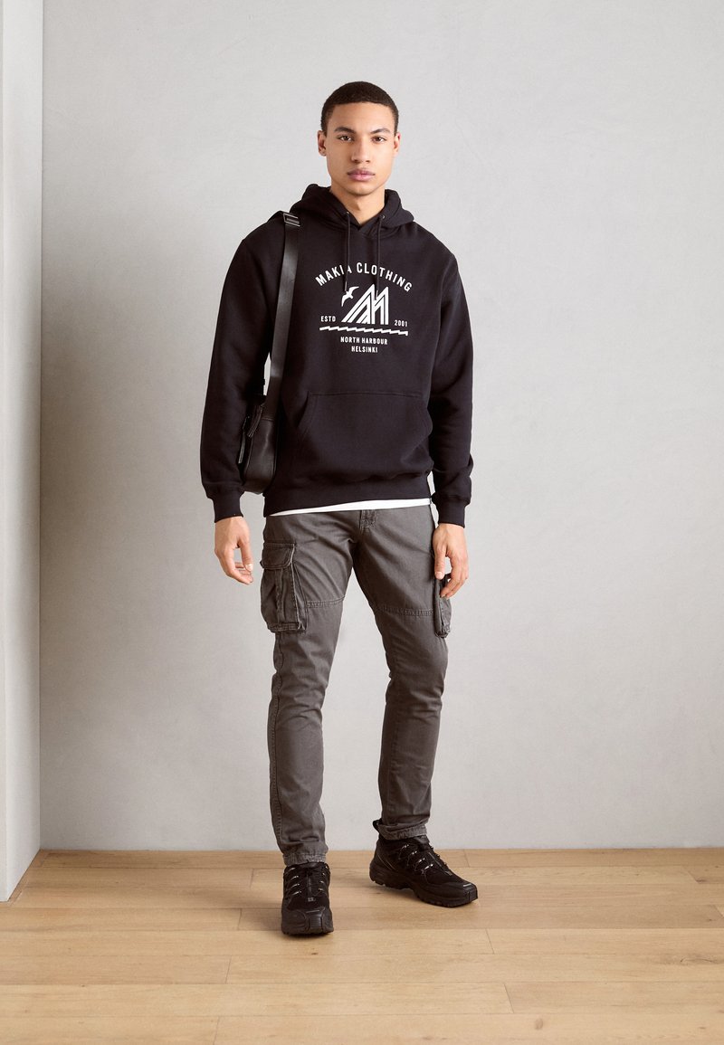 Sudadera negra con capucha y logo gráfico blanco, pantalones cargo grises con bolsillos laterales y zapatillas negras. Suelo de madera clara y pared neutra.