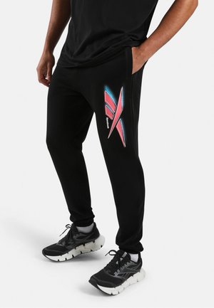 Personne portant un pantalon de jogging noir avec un logo Reebok rouge et bleu sur la cuisse, des baskets noires et un t-shirt noir, debout avec une main dans la poche.