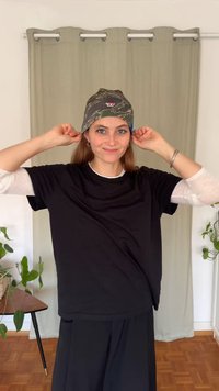 Kšiltovka s maskáčovým vzorem a logem, nošená s černým tričkem přes bílé tričko s dlouhým rukávem, na neutrálním pozadí záclony.