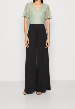 Žena v hrdzavě zeleném krajkovém topu s krátkými volánovými rukávy, v kombinaci s černými kalhotami s širokými nohavicemi a vysokým pasem a černými sandály.