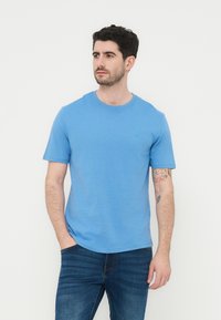 Άνδρας με κοντά σκούρα μαλλιά, που φοράει ανοιχτό μπλε μπλουζάκι και σκούρο τζιν, στέκεται με το ένα χέρι στην τσέπη, κοιτώντας προς το πλάι.