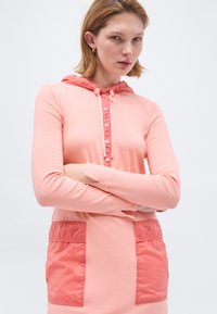 Νεαρή γυναίκα με κοντά ξανθά μαλλιά που φοράει μακρυμάνικο ροζ φόρεμα με κουκούλα, μεγάλα μπροστινά τσέπες και κουμπιά.