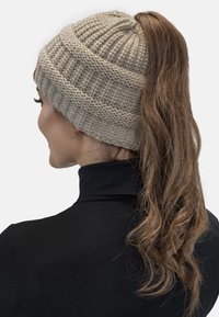 Bonnet en tricot beige avec texture côtelée, conçu pour accueillir une queue de cheval, avec un dessus arrondi et des détails de couture subtils.