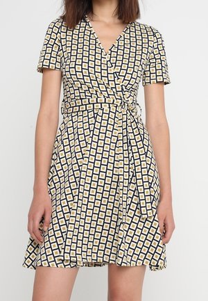 Robe portefeuille à manches courtes avec motif à carreaux noirs, blancs et jaunes et détail de nœud sur le côté, portée par une personne debout.