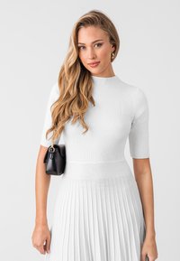 Haut en tricot côtelé blanc avec un col haut et des manches courtes, associé à une jupe plissée blanche. Sac à main noir avec des accents en métal doré.