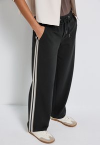 Personne portant un pantalon large noir avec des rayures blanches sur les côtés, un blazer beige et des sneakers blanches, se tenant les mains dans les poches.