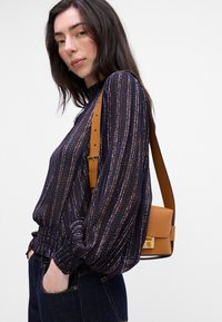 Blusa blu marina con righe dorate, polsini arricciati, abbinata a una borsa a tracolla marrone con una chiusura dorata. Jeans in denim scuro completano il look.