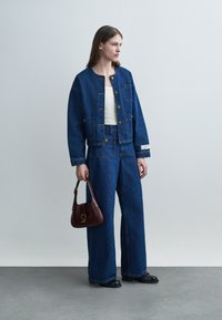 Kvinne iført mørkeblå denimjakke og vide bukser, hvit topp, svarte sko, holder en mørkerød håndveske, står mot en ensfarget grå bakgrunn.