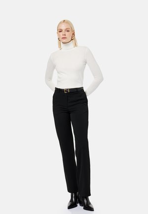 Frau steht mit den Händen hinter dem Rücken, trägt einen weißen Rollkragenpullover, schwarze Hose mit Gürtel, schwarze Stiefeletten und goldene Creolen.