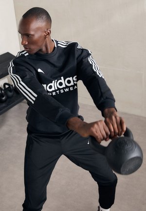 Felpa sportiva nera Adidas con logo bianco, abbinata a pantaloni neri. Tieni in mano un kettlebell nero, impegnato in un allenamento.