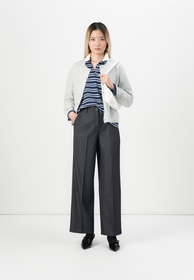 Donna che indossa un cardigan grigio, una maglietta a righe blu navy, pantaloni larghi scuri, scarpe nere e tiene una borsa a tracolla bianca su uno sfondo bianco.