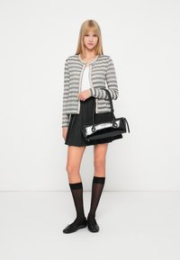 Stripet grå cardigan, hvit topp, svart plisserte skjørt, svart håndveske, knelengde sokker og svarte leiligheter på en nøytral bakgrunn.