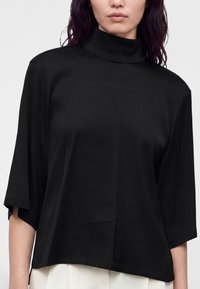 Femme portant un haut noir ample à col montant avec des manches aux coudes et un pantalon blanc, vue des épaules aux hanches.