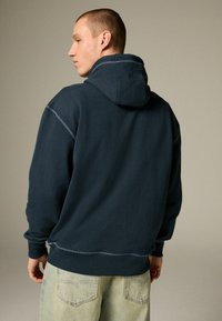 Sudadera oscura de color marino con cordón, que presenta detalles de costura visibles. Textura de tela suave con un ajuste relajado, combinada con jeans de mezclilla claros.